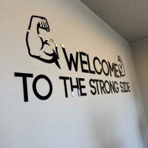 שילוט אקרילי שחור באורך 2 מטר עם הכתובת WELCOME TO THE STRONG SIDE ואיור זרוע כפופה על קיר לבן בחדר כושר