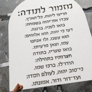 לוח מזמור לתודה מעץ בצביעה לבנה עם אותיות בולטות בגילוף, מידה 75/120 ס"מ, מוחזק על רקע קיר בטון