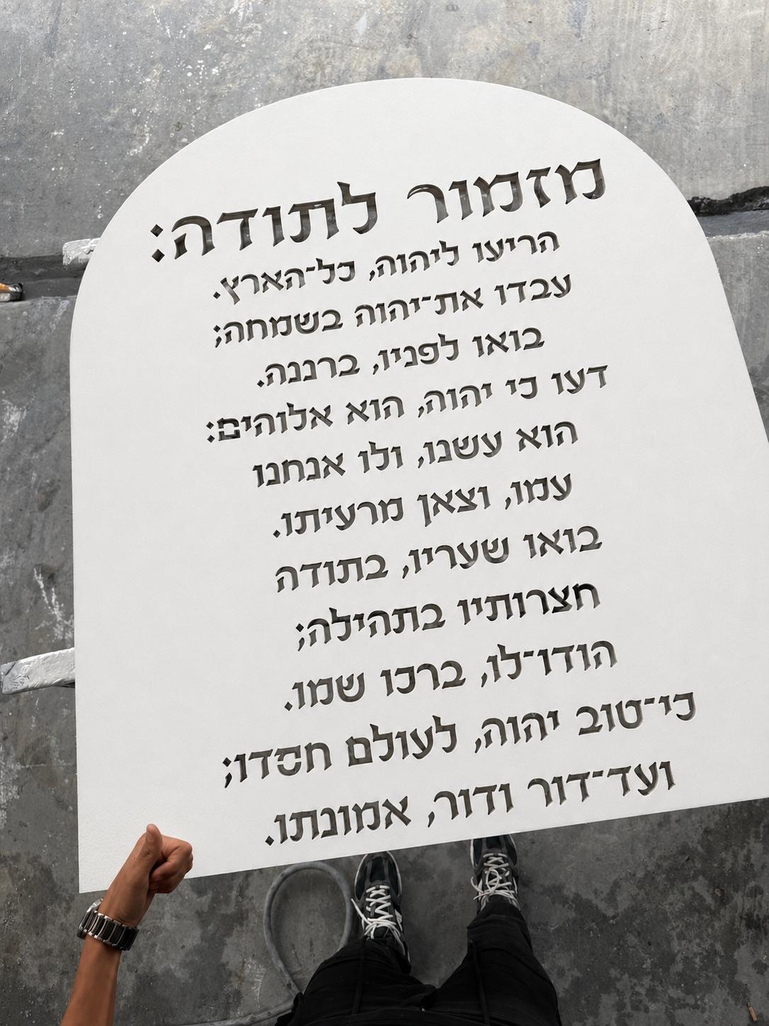 לוח מזמור לתודה מעץ בצביעה לבנה עם אותיות בולטות בגילוף, מידה 75/120 ס"מ, מוחזק על רקע קיר בטון