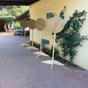ארבעה מעמדי עץ עגולים בגוון טבעי בהיר עם בסיס רגליים, ניצבים בשורה על משטח אבן לצד קיר בכניסה לאירוע