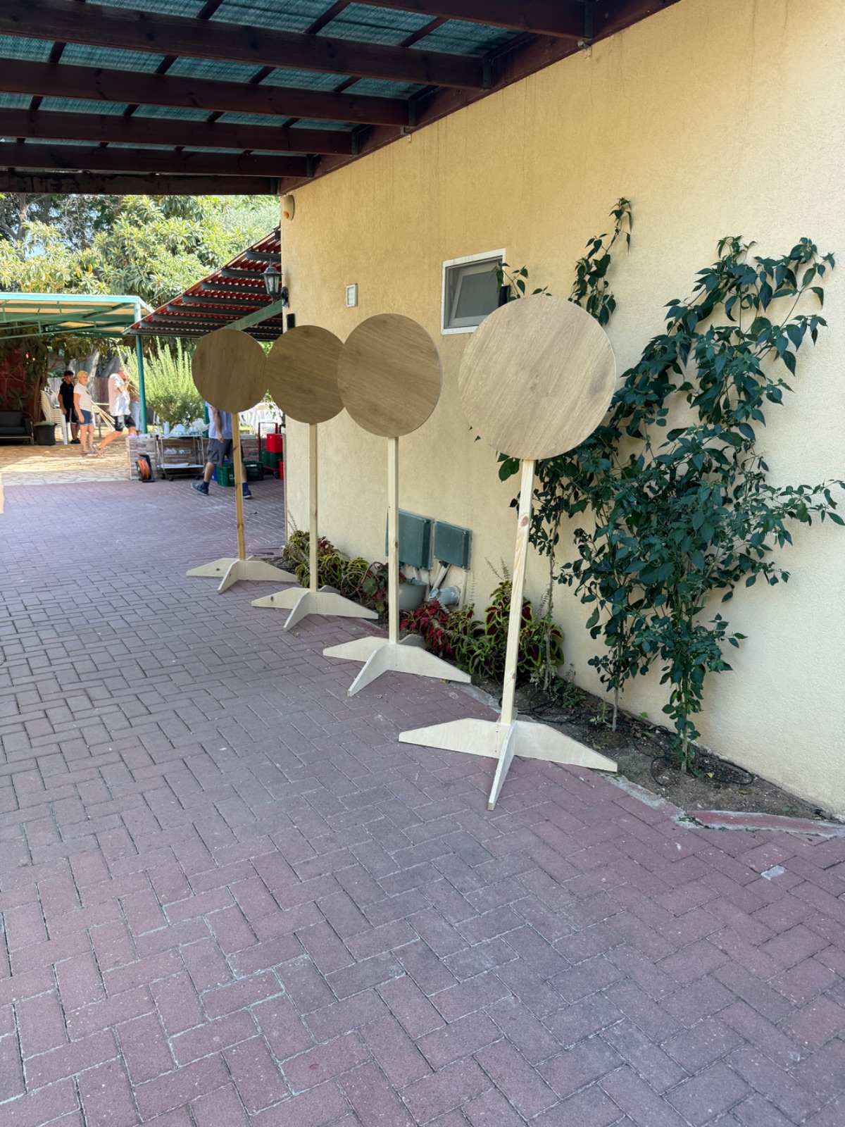 ארבעה מעמדי עץ עגולים בגוון טבעי בהיר עם בסיס רגליים, ניצבים בשורה על משטח אבן לצד קיר בכניסה לאירוע