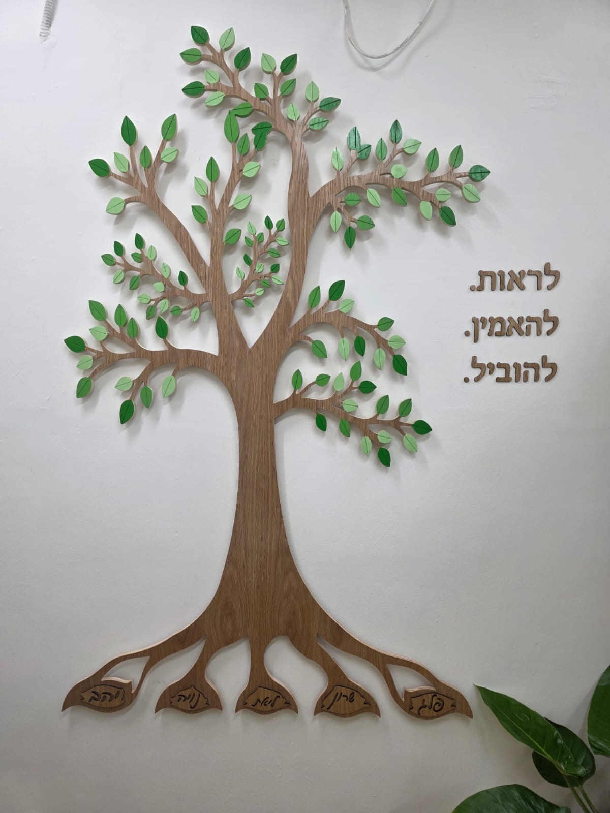 עץ קיר תלת־ממדי מעץ עם עלים ירוקים ושמות מגולפים בשורשים, מותקן על קיר לבן לצד הכיתוב לראות להאמין להוביל